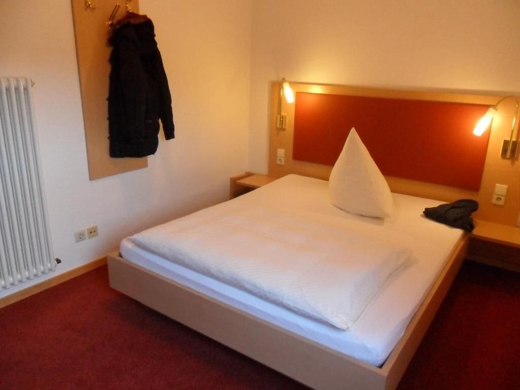 Habitación de hotel con una cama individual, cabecera con iluminación integrada, mesita de noche y perchero.
