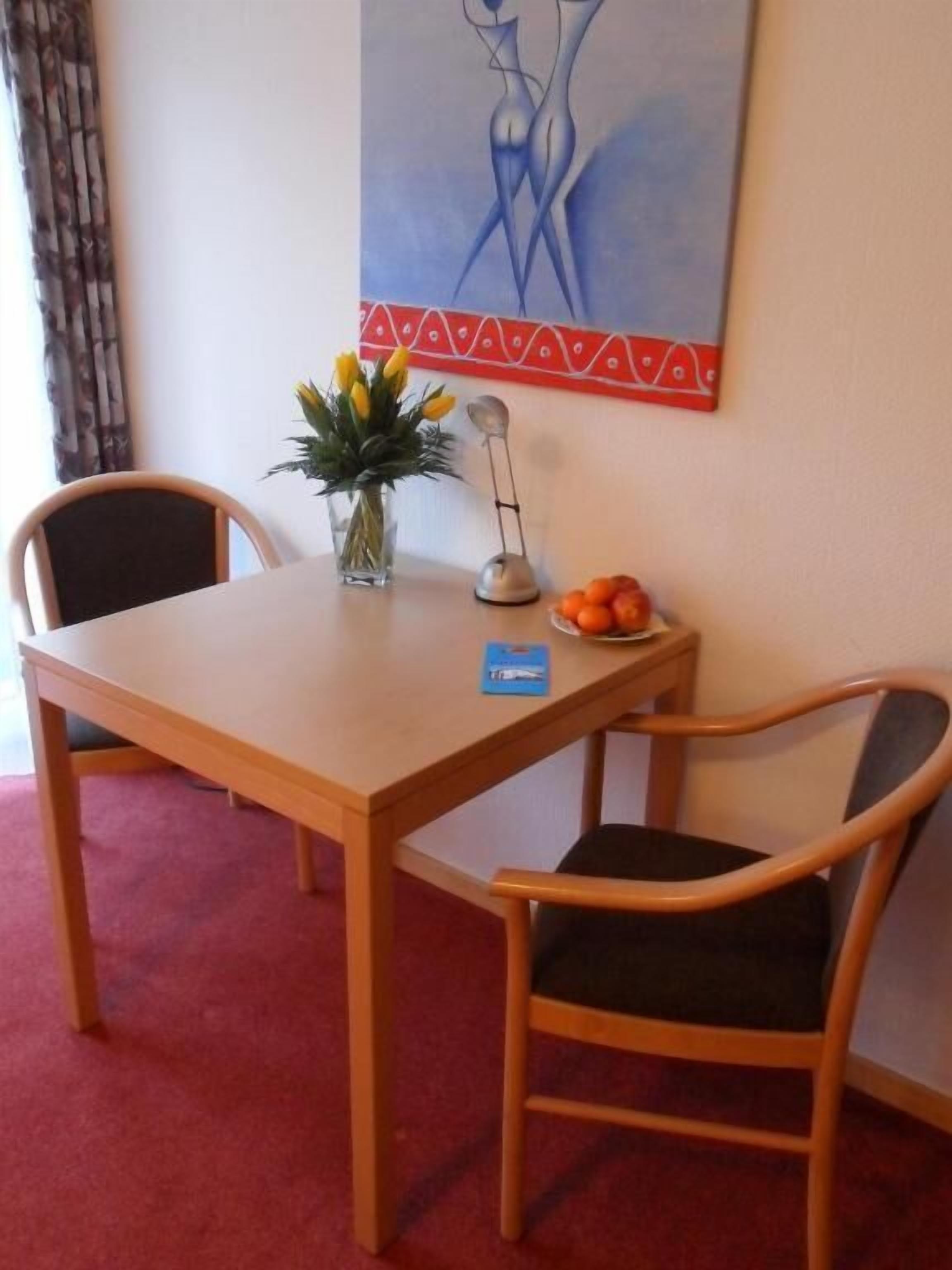 A dining area with a wooden table and two chairs, a vase with flowers, a fruit plate, and a painting on the wall.