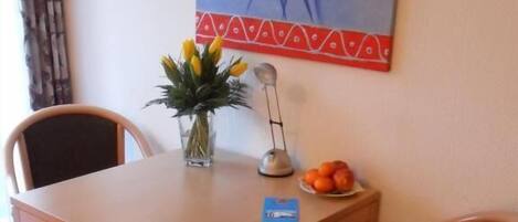A dining area with a wooden table and two chairs, a vase with flowers, a fruit plate, and a painting on the wall.