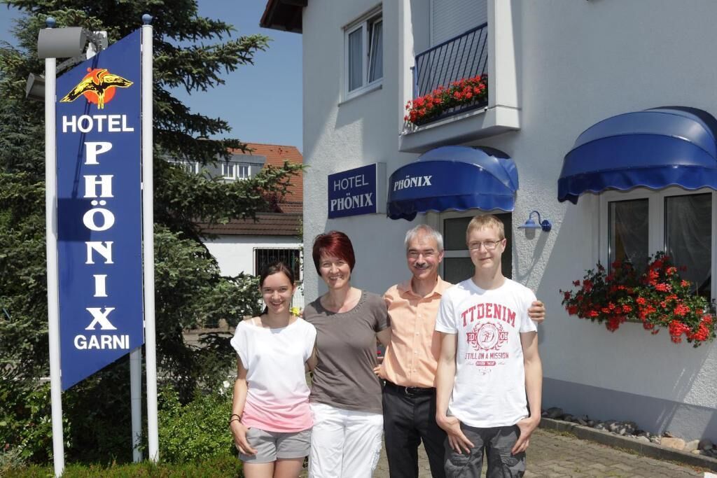 A group of people standing in front of Hotel Phönix Garnier, with a blue sign and a building with a balcony and flower boxes.