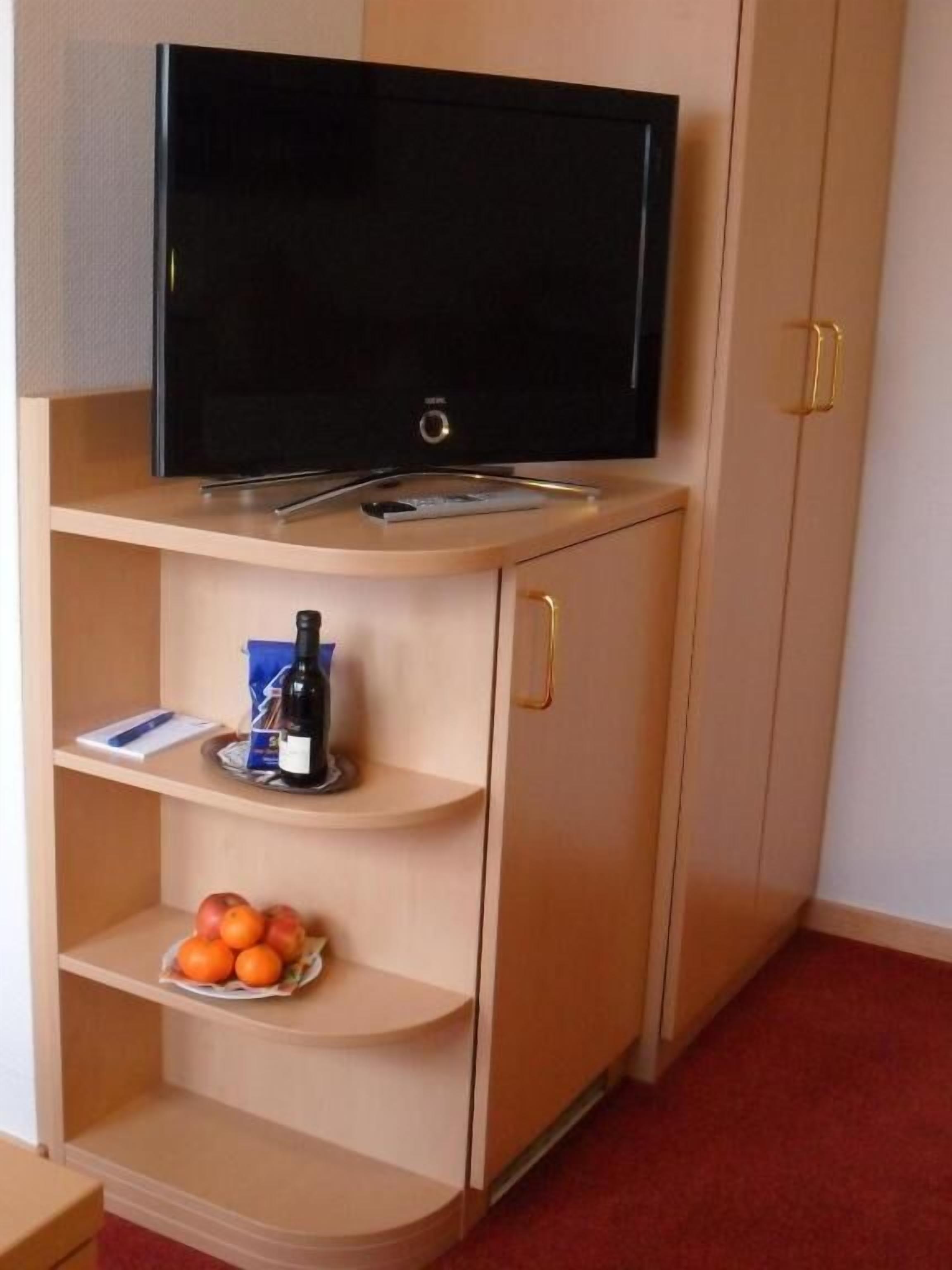 A room with a television on a shelf, a bottle of wine, and a bowl of oranges on a lower shelf.