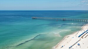 A beach with a long pier extending into the ocean, clear turquoise water, and a sandy shore with umbrellas and beach chairs.