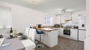 A modern kitchen with white cabinetry, a central island with bar stools, a built-in oven, and a microwave. - Broads Escape/ - 3 Bedroom House in Oulton Broads (Lowestoft)