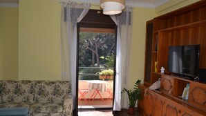 A living room with a sofa, a TV on a wooden cabinet, a balcony with a table and chairs, and a view of trees and plants outside. - Cozy apartment a stone's throw from the historic center of Rome (Roma)