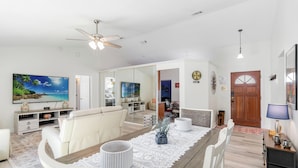 A spacious living area with a wooden dining table, white chairs, a ceiling fan, and a large TV.