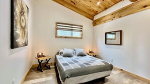 A bedroom with a wooden ceiling, a bed with a grey and white checkered bedspread, two bedside tables with lamps, a window with blinds, and a wall-mounted mirror.