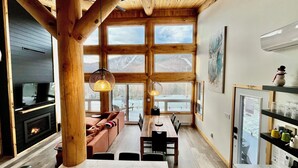 A cozy cabin interior with a fireplace, wooden beams, large windows, a dining area, and a kitchen with shelves.