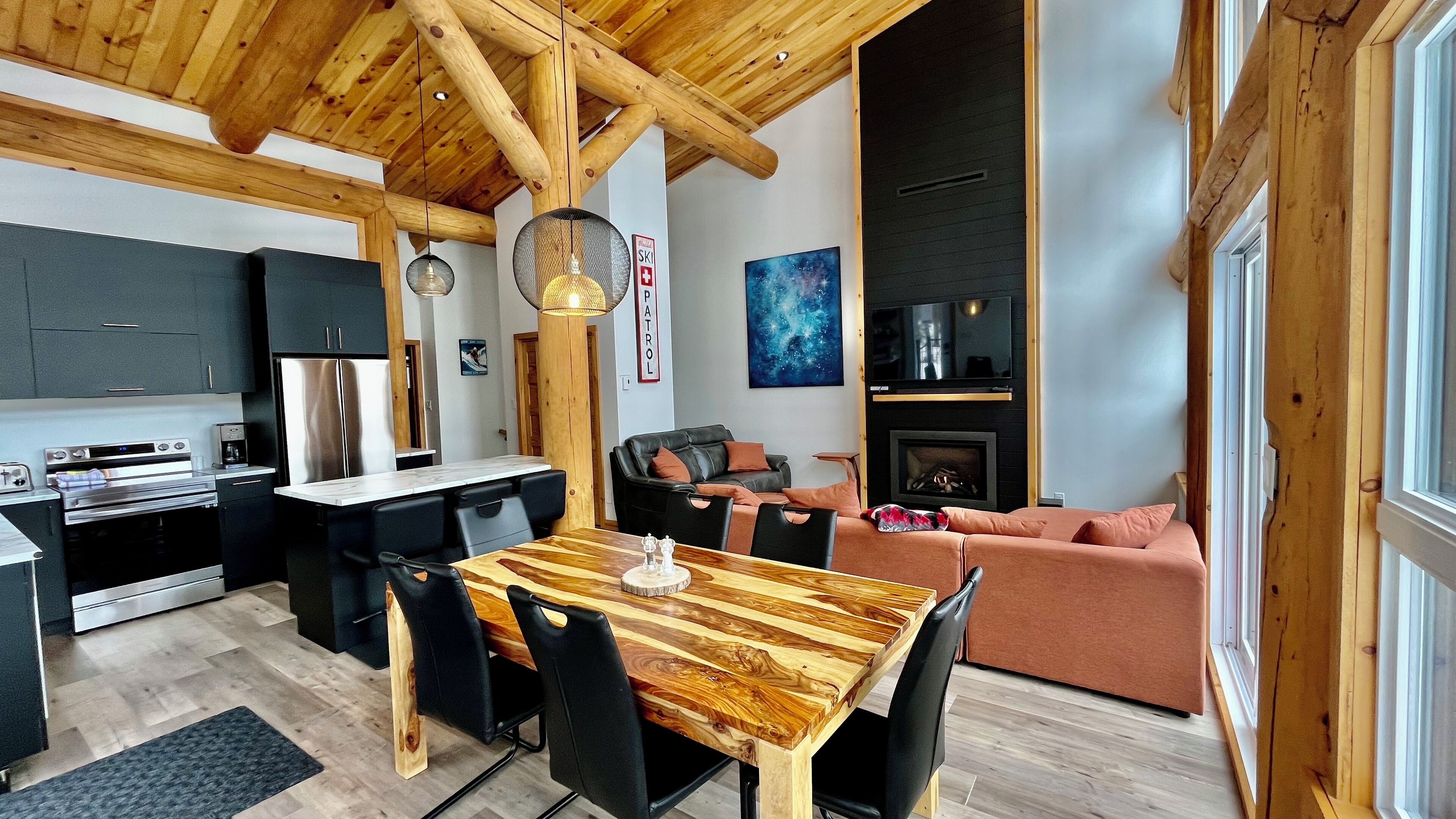 Una cuina moderna amb forn integrat i nevera, una zona de menjador amb taula i cadires de fusta, un saló amb sofà i llar de foc, i un gran finestral que proporciona llum natural.