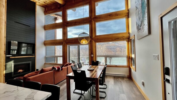 A spacious living area with a fireplace, a dining table with chairs, and large windows offering a view of the outdoors.