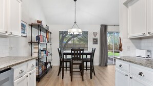 Una cocina con gabinetes blancos, encimera de granito, una mesa de comedor con sillas, una lámpara de araña y una ventana con persianas.