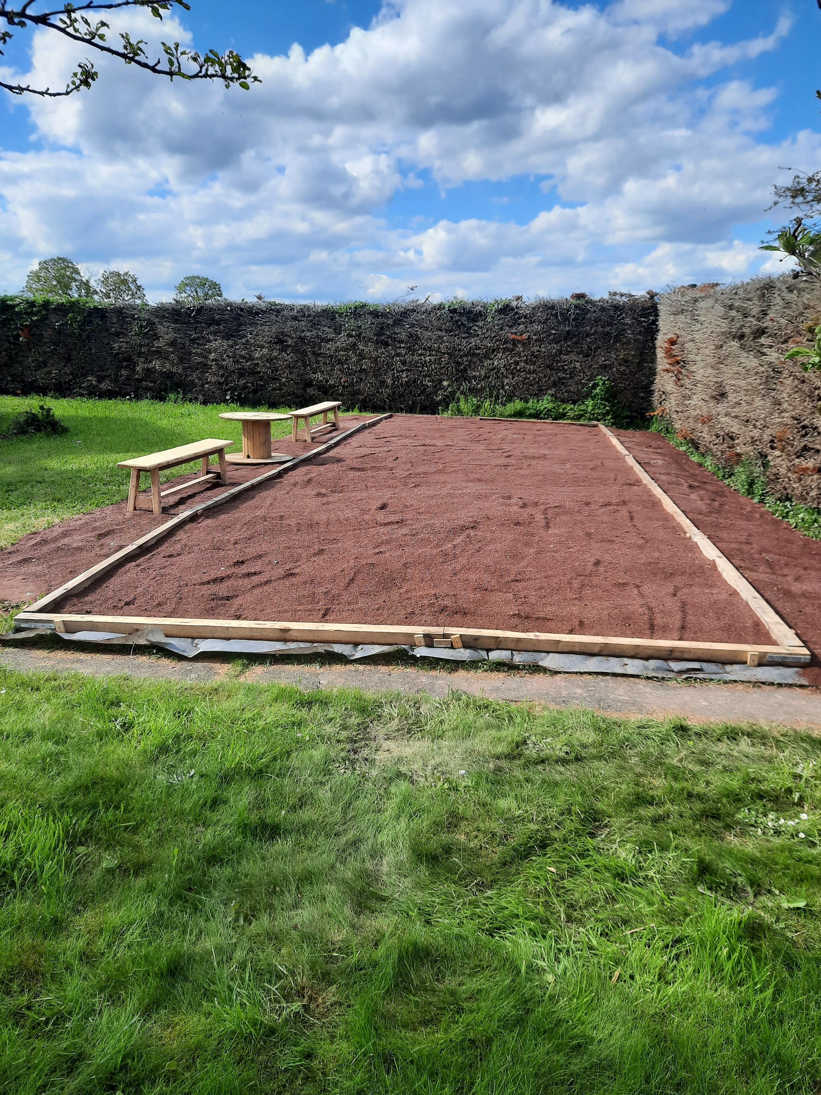 A garden with a raised red soil bed, two wooden benches, and a hedge in the background.