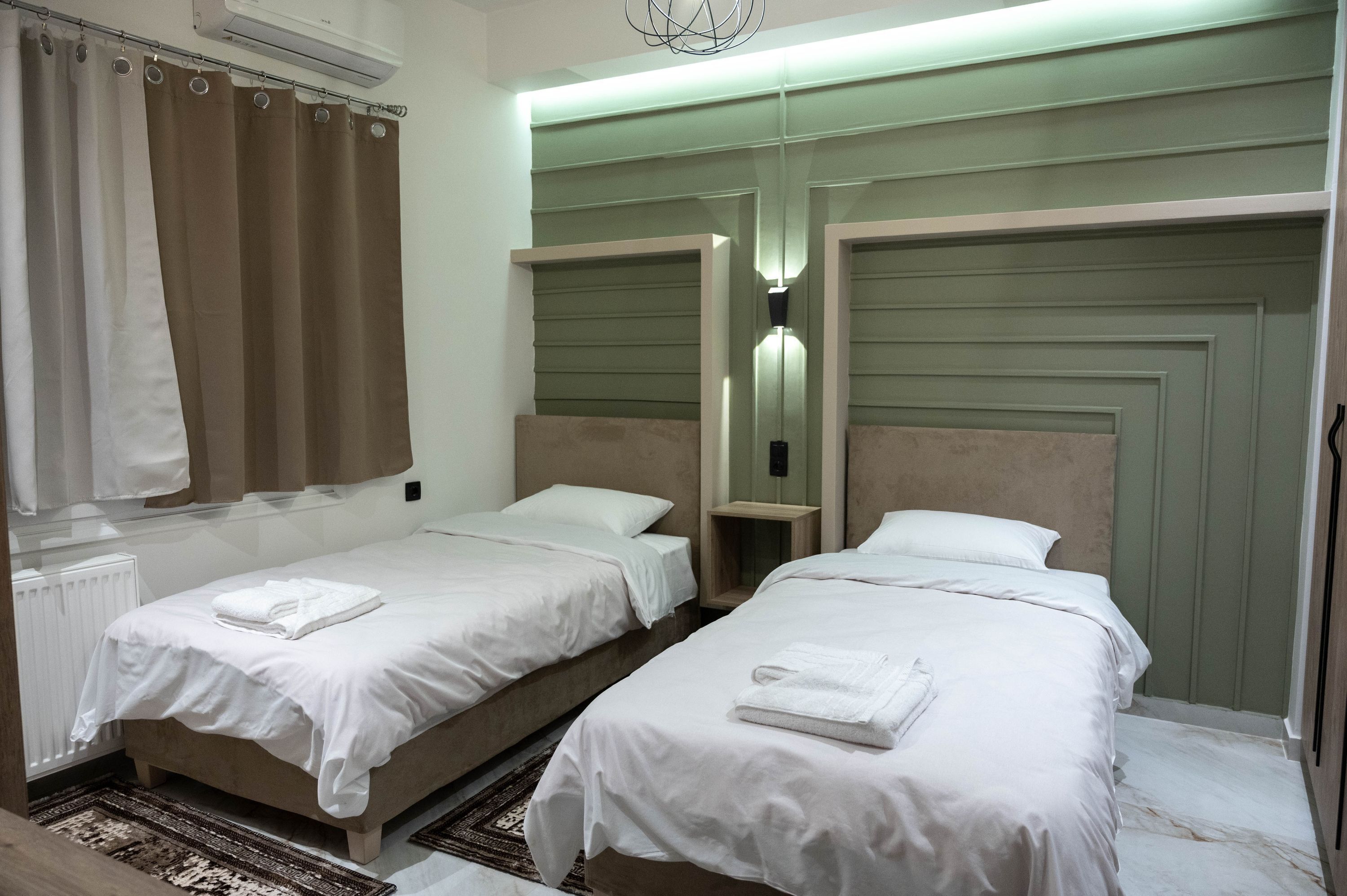A hotel room with two single beds, a radiator, a curtain, and a wall-mounted light fixture.