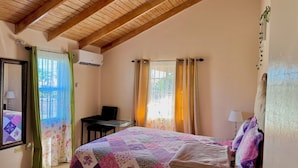 Ein Schlafzimmer mit Holzbalkendecke, einem Bett mit bunter Steppdecke, einem Schreibtisch und einem Stuhl.