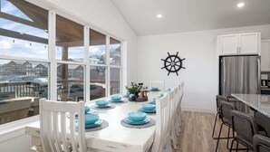Een eetgedeelte met een witte tafel gedekt voor zes personen, een stuurrad aan de muur en een uitzicht op huizen door grote ramen.