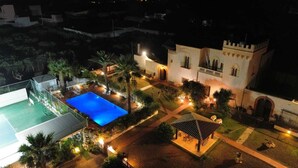 Una gran piscina envoltada per un pati ben il·luminat amb un edifici semblant a un castell, una pista de tennis i un porxo.