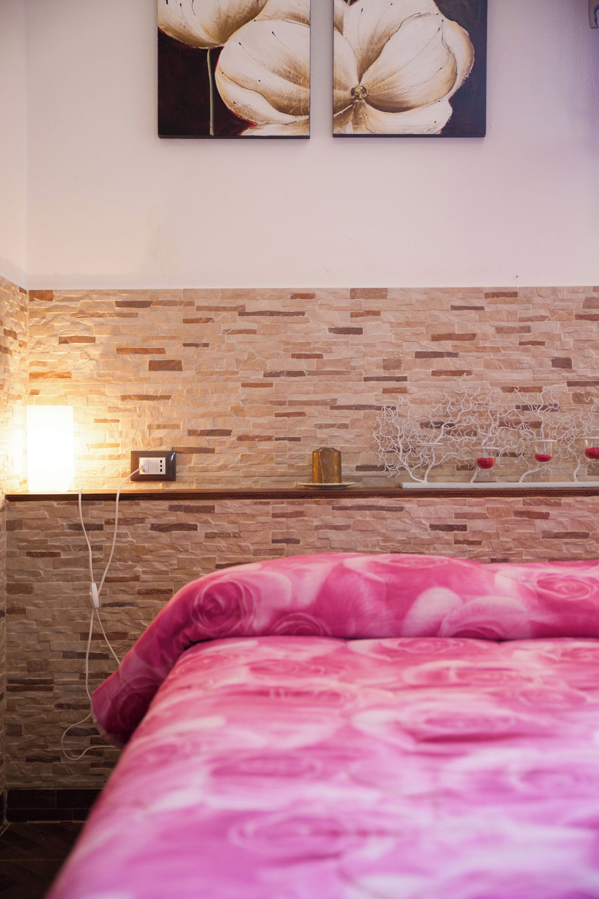 Quarto com parede em tijolos, uma cama com colcha floral rosa e dois quadros de flores pendurados na parede.