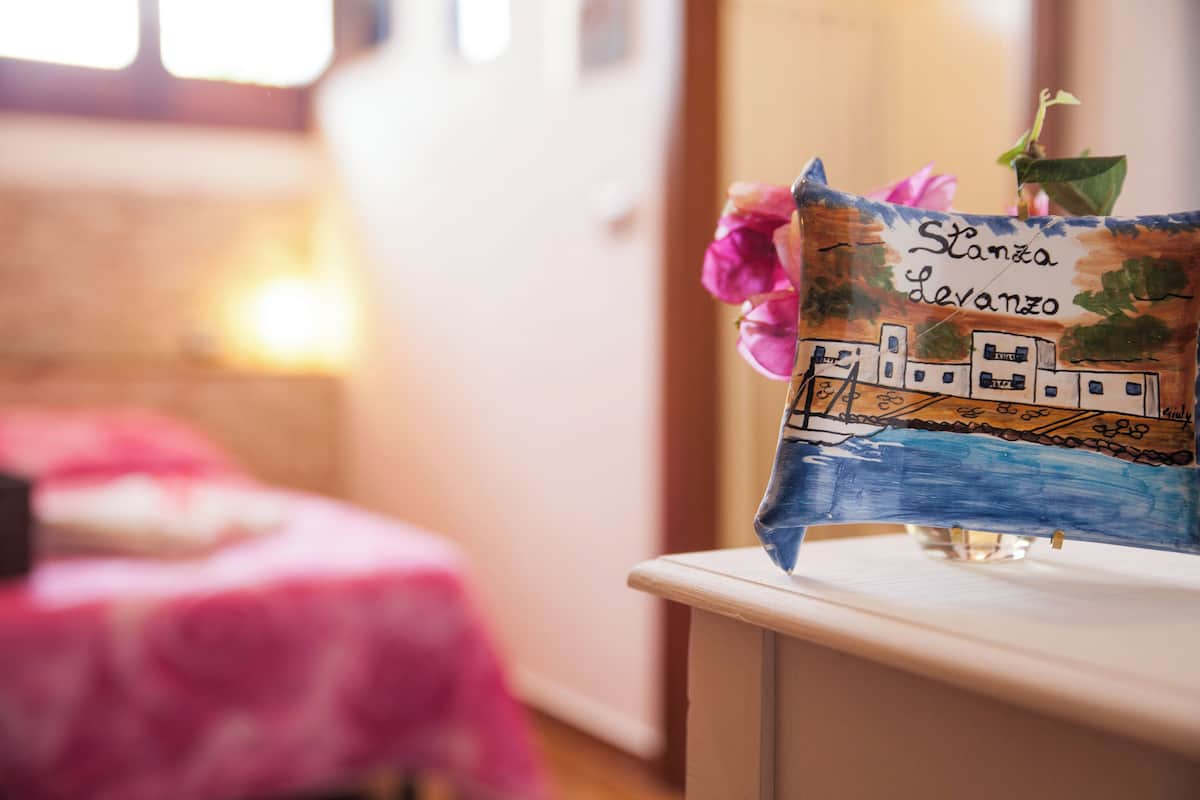 Un coussin peint à la main, orné d’un motif représentant un paysage urbain et d’une fleur rose, ainsi qu’un vase en verre transparent posé sur une commode blanche.