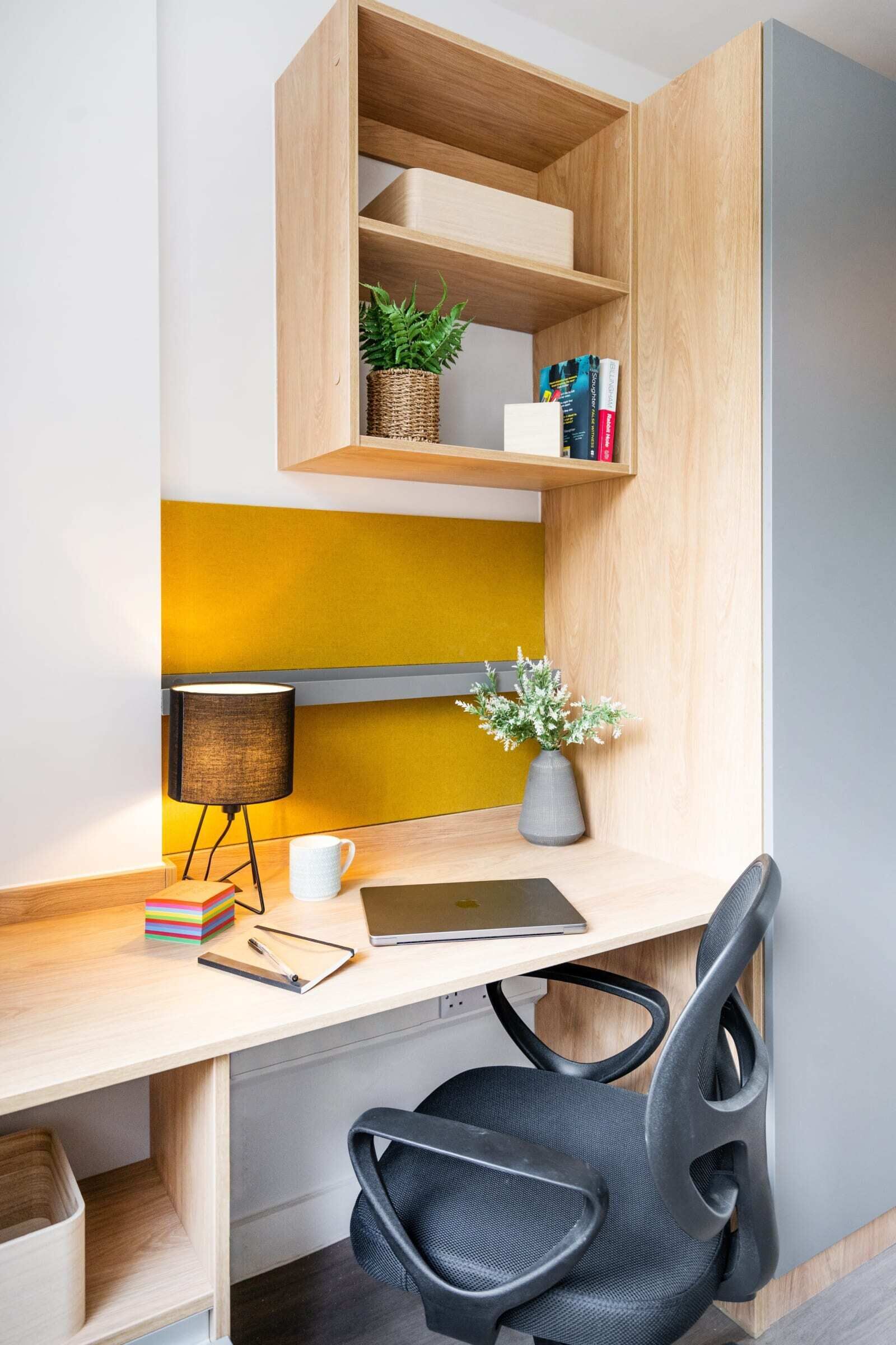 A modern office desk with a laptop, a lamp, and a chair. There are shelves above the desk with plants and books.