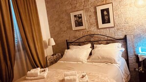 Ein Hotelzimmer mit Steinwand, einem Bett mit weißen Leinen, zwei gerahmten Bildern an der Wand und einem Fenster mit Vorhängen.