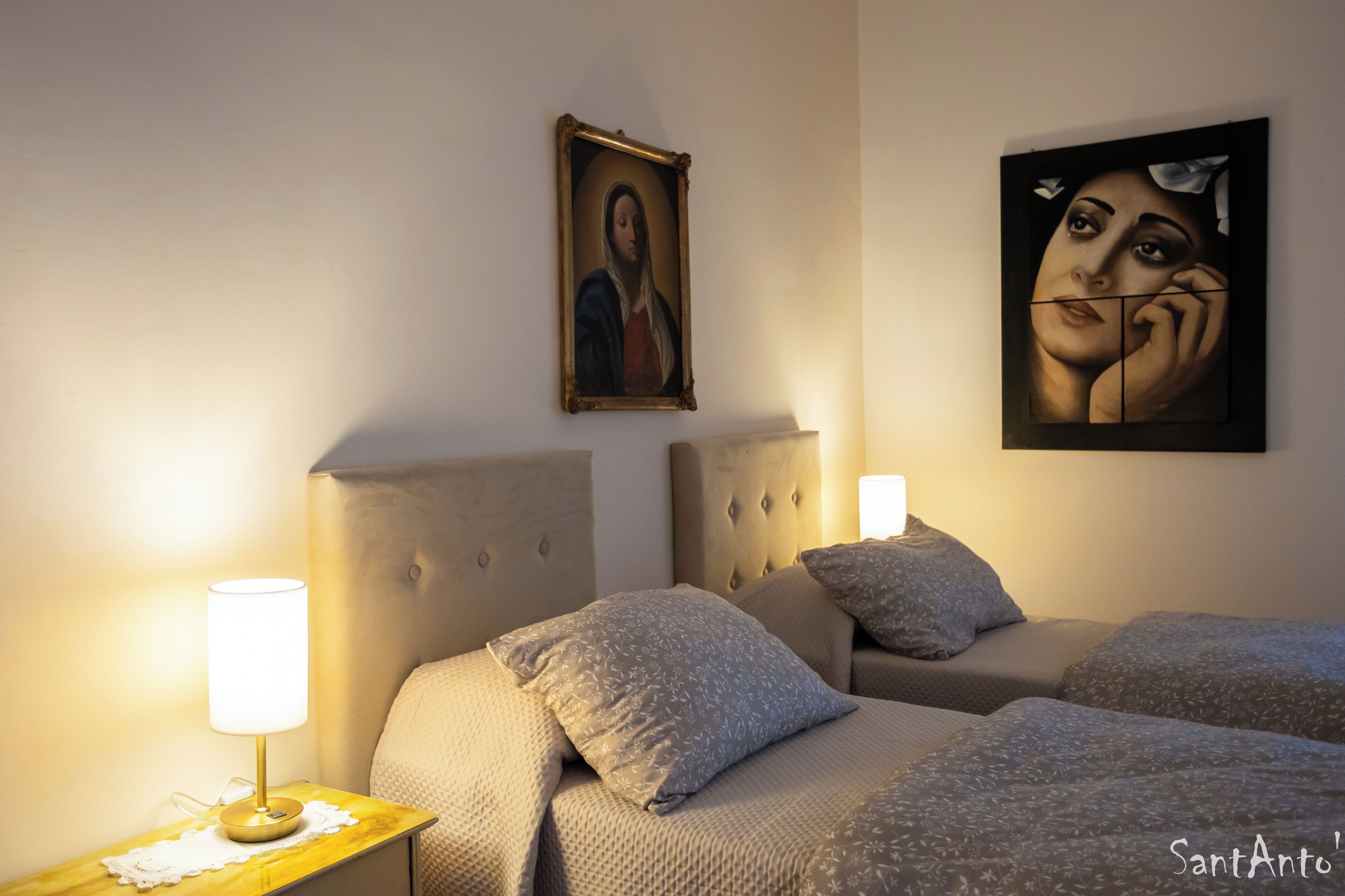 Una camera d'albergo con due letti, un comodino, una lampada e due quadri appesi al muro.
