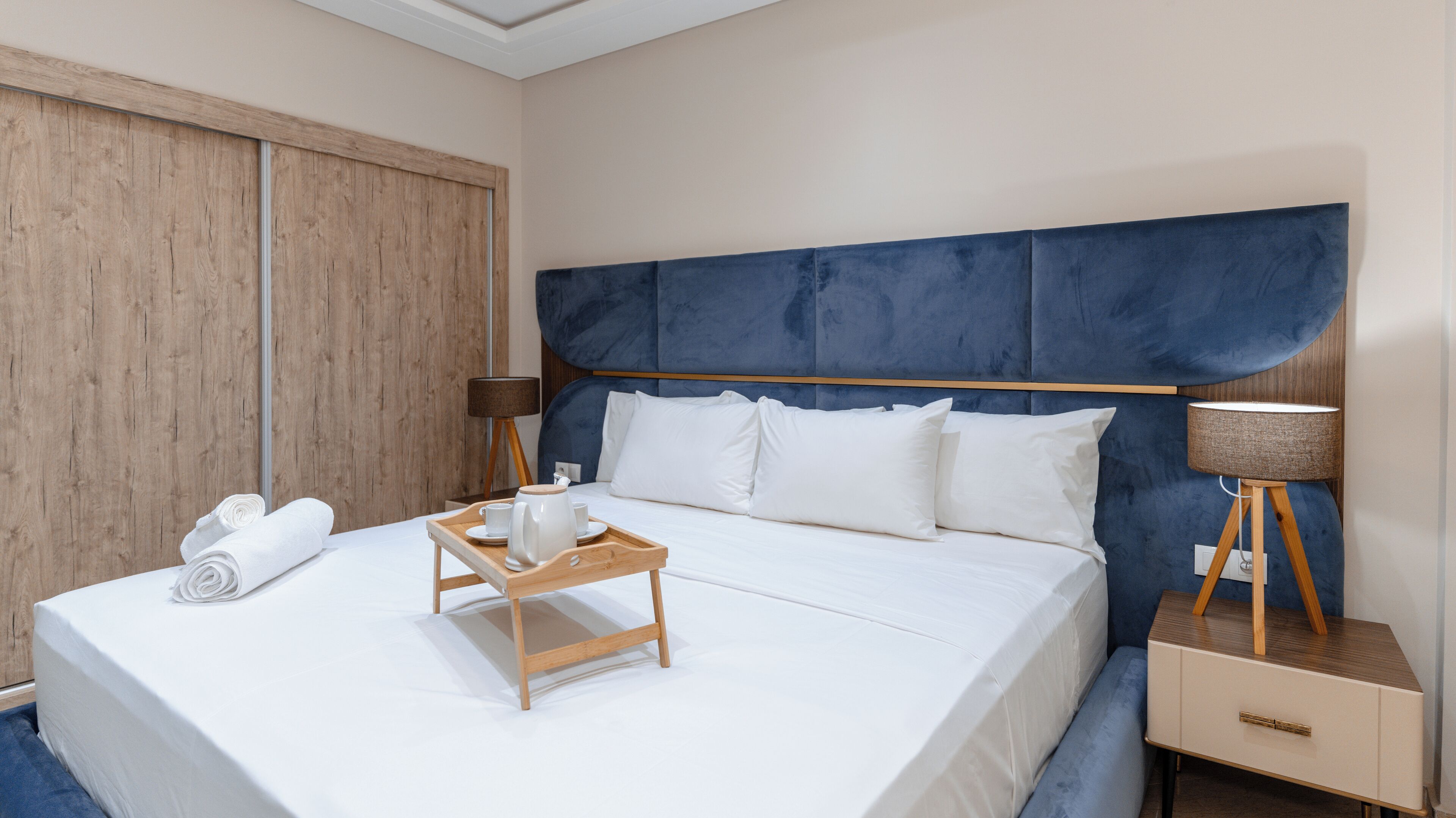 Un lit bien fait, avec du linge de lit blanc, une table de chevet en bois sur laquelle se trouve une lampe, et un plateau avec une bouilloire et des tasses.