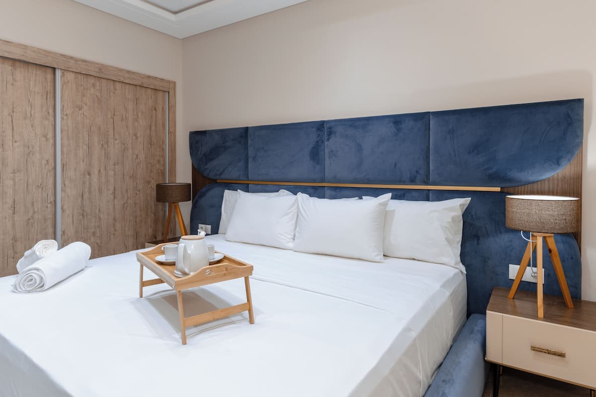 A neatly made bed with white linens, a wooden bedside table with a lamp, and a tray with a kettle and cups.