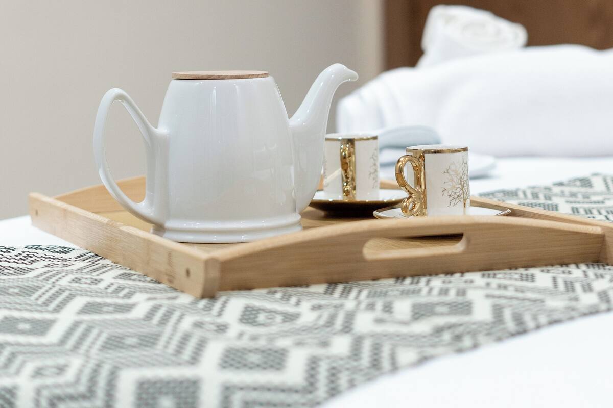 Un plateau en bois sur lequel reposent une théière blanche et deux tasses, le tout posé sur un lit recouvert d’une literie à motifs.