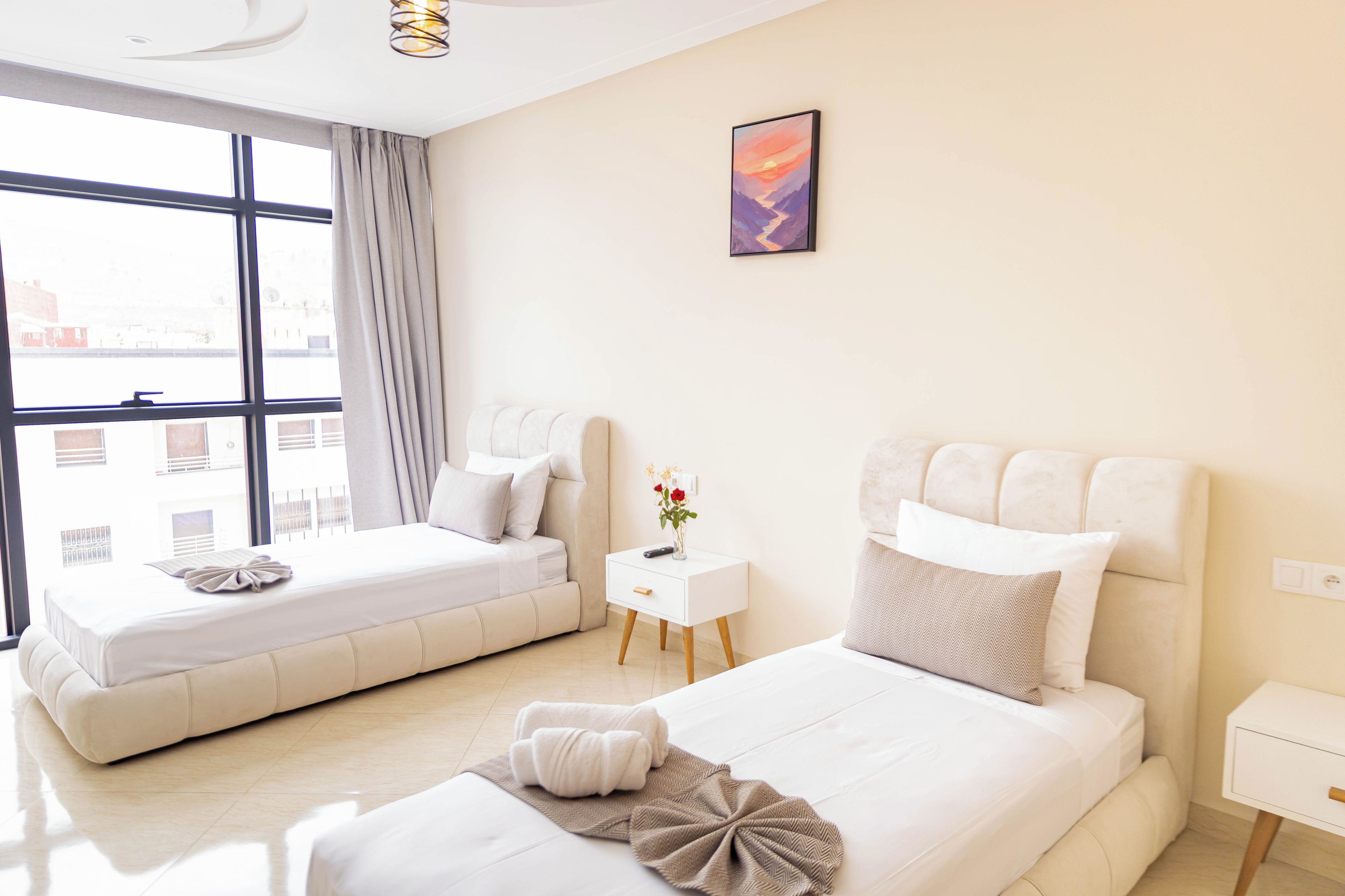 A hotel room with two beds, a small bedside table, a wall-mounted artwork, and a view of the city through large windows.