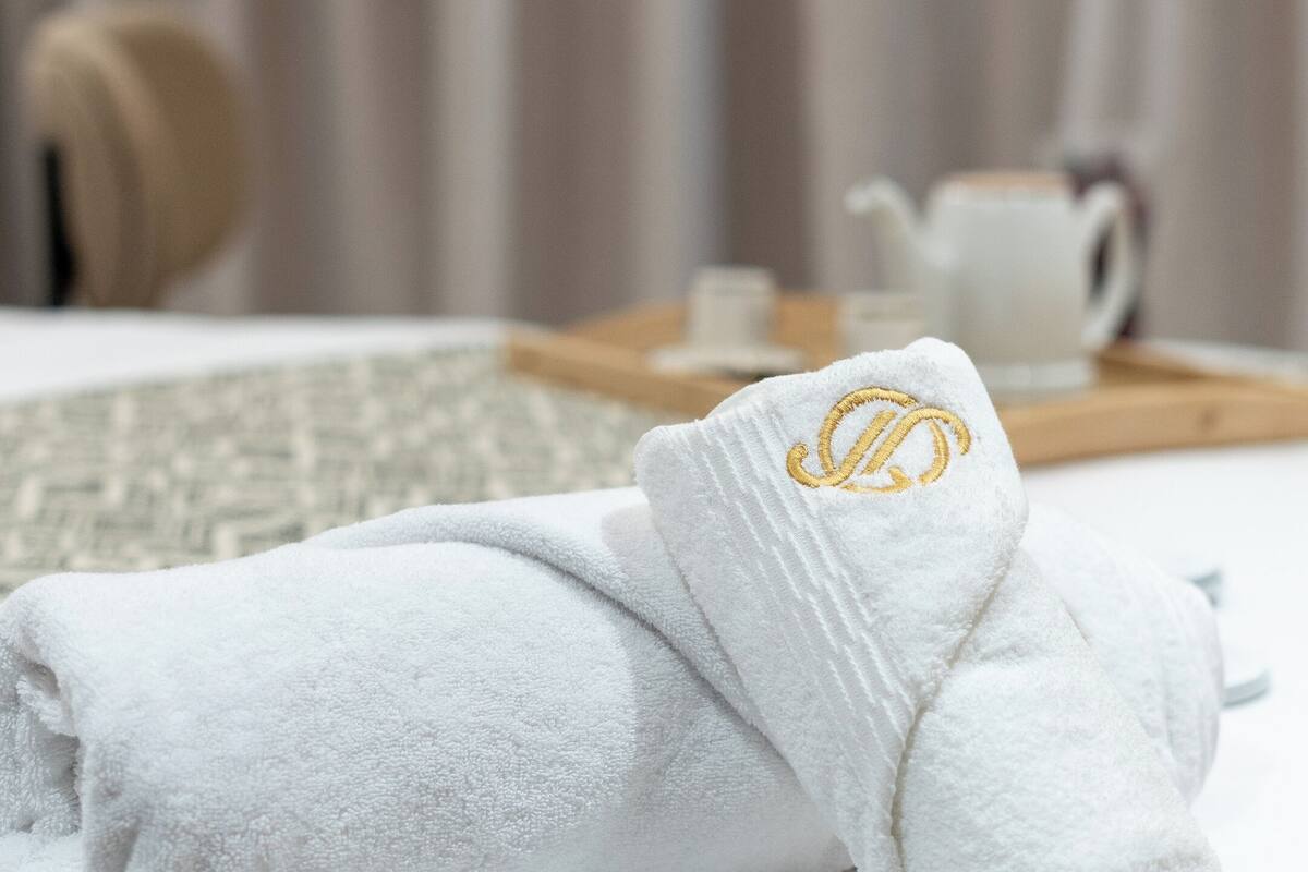Une serviette blanche, soigneusement pliée, ornée d’un écusson doré, est posée sur un lit recouvert d’une couverture blanche.