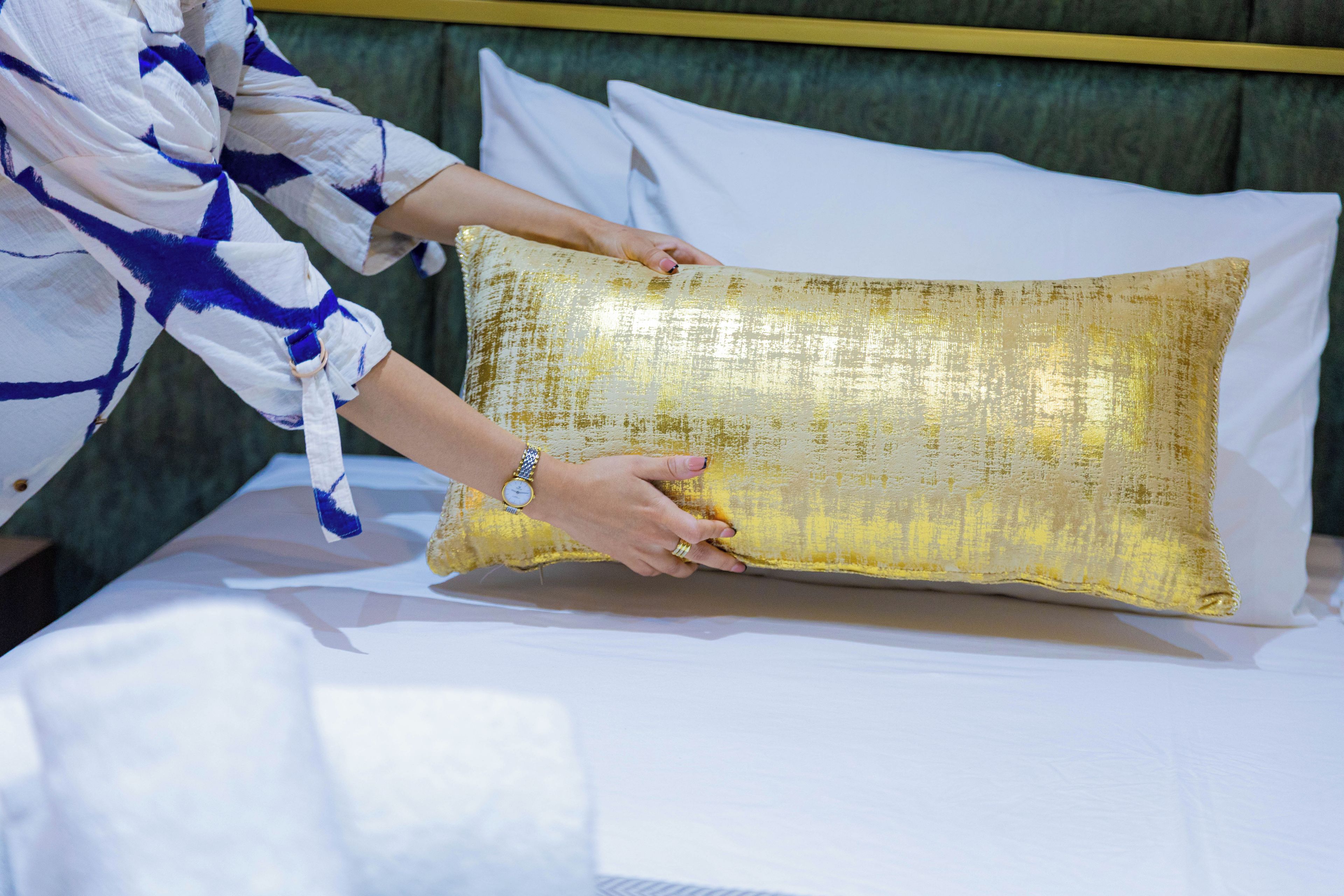 A person placing a gold textured pillow on a bed with white sheets.