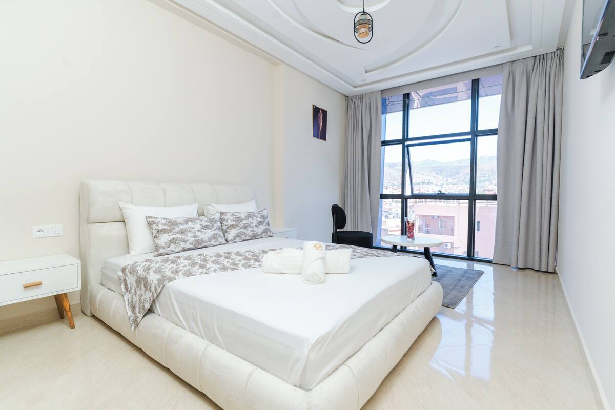 Une chambre d’hôtel moderne, dotée d’un grand lit, d’une petite table, d’une chaise et d’une grande fenêtre donnant sur la ville.