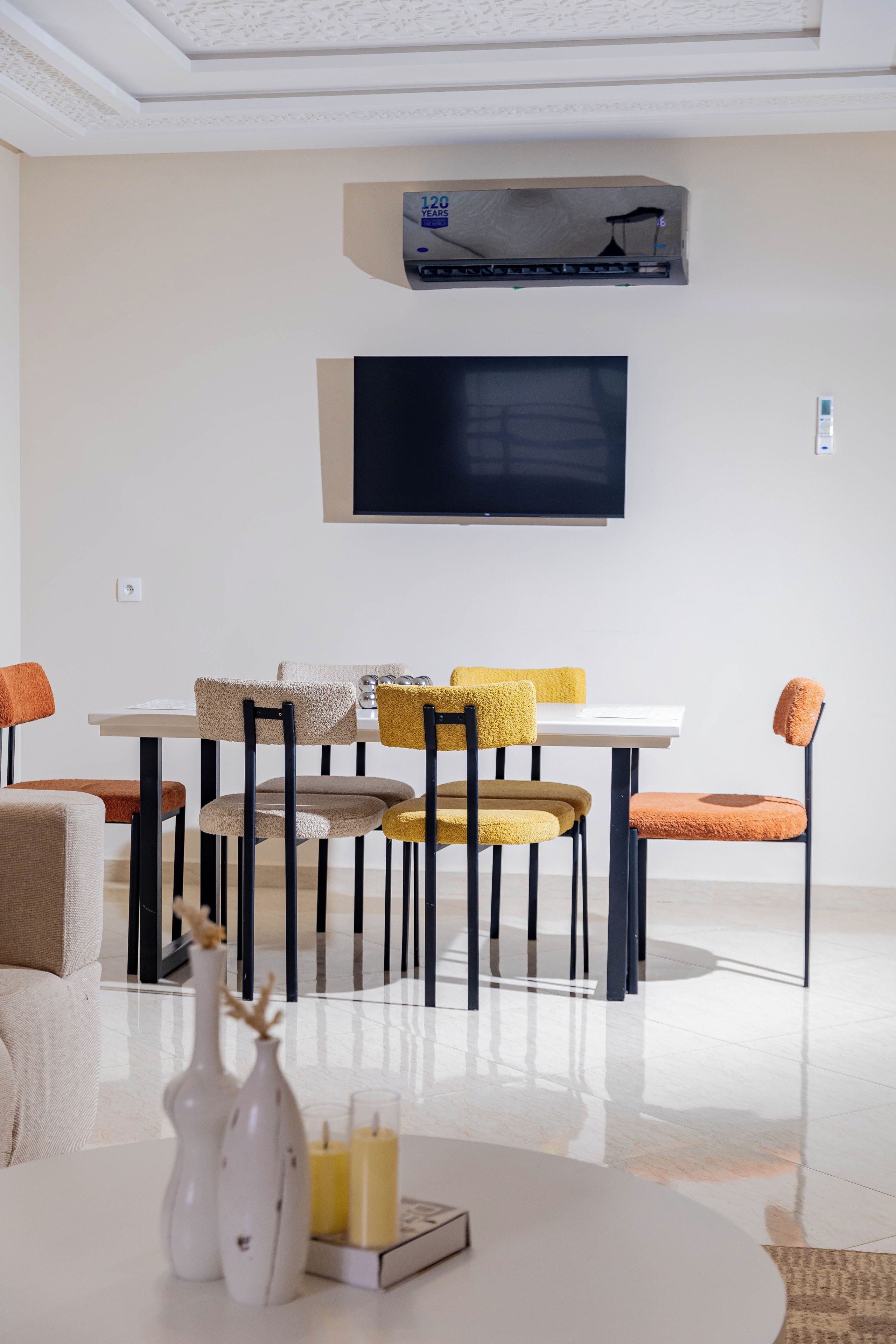 A modern dining area with a white table, multiple chairs, a flat-screen TV mounted on the wall, and a ceiling air conditioning unit.