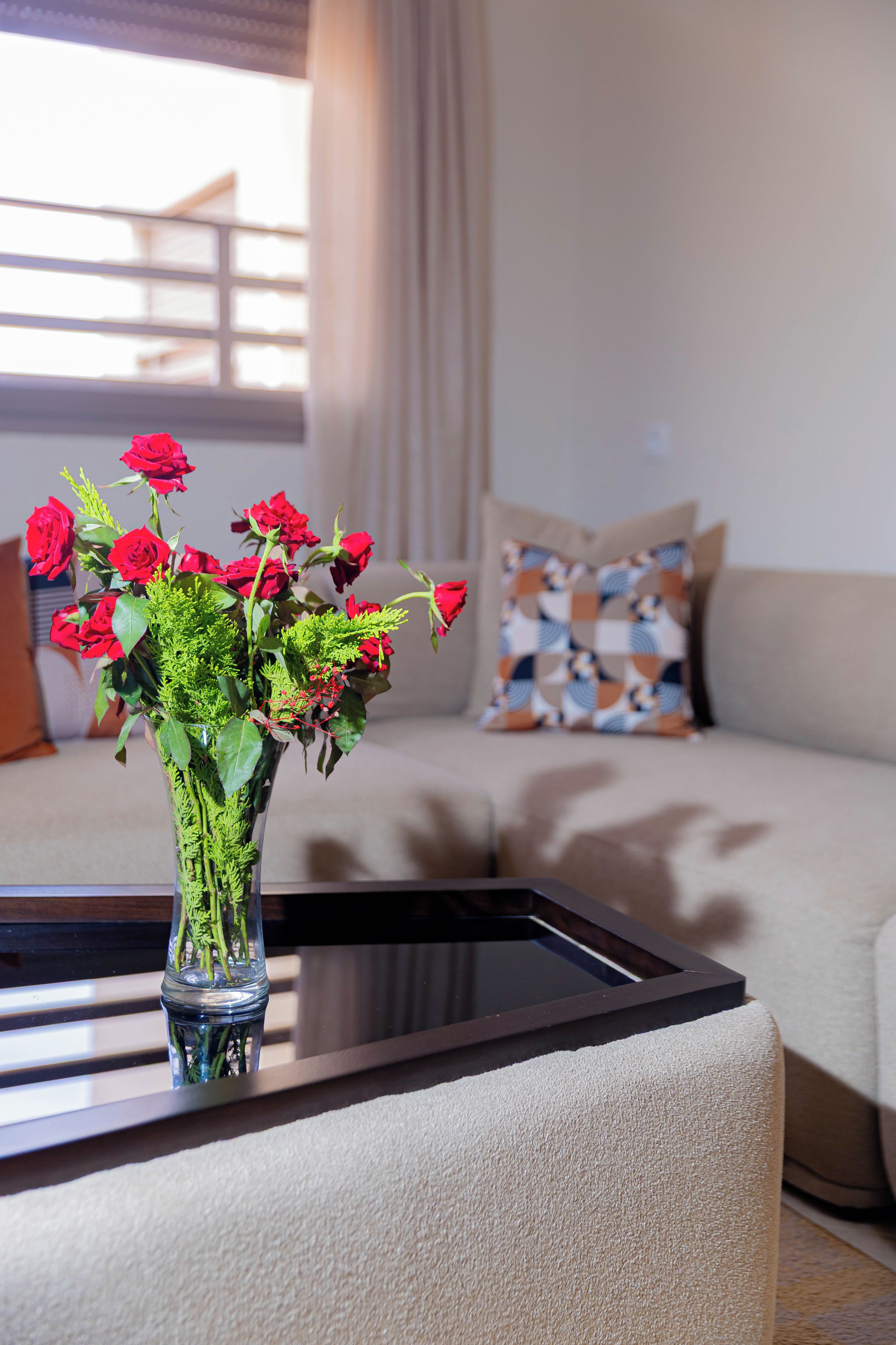 A living room with a beige sofa, a patterned pillow, and a vase with red flowers on a glass table.