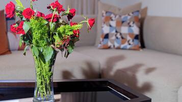 A living room with a beige sofa, a patterned pillow, and a vase with red flowers on a glass table.