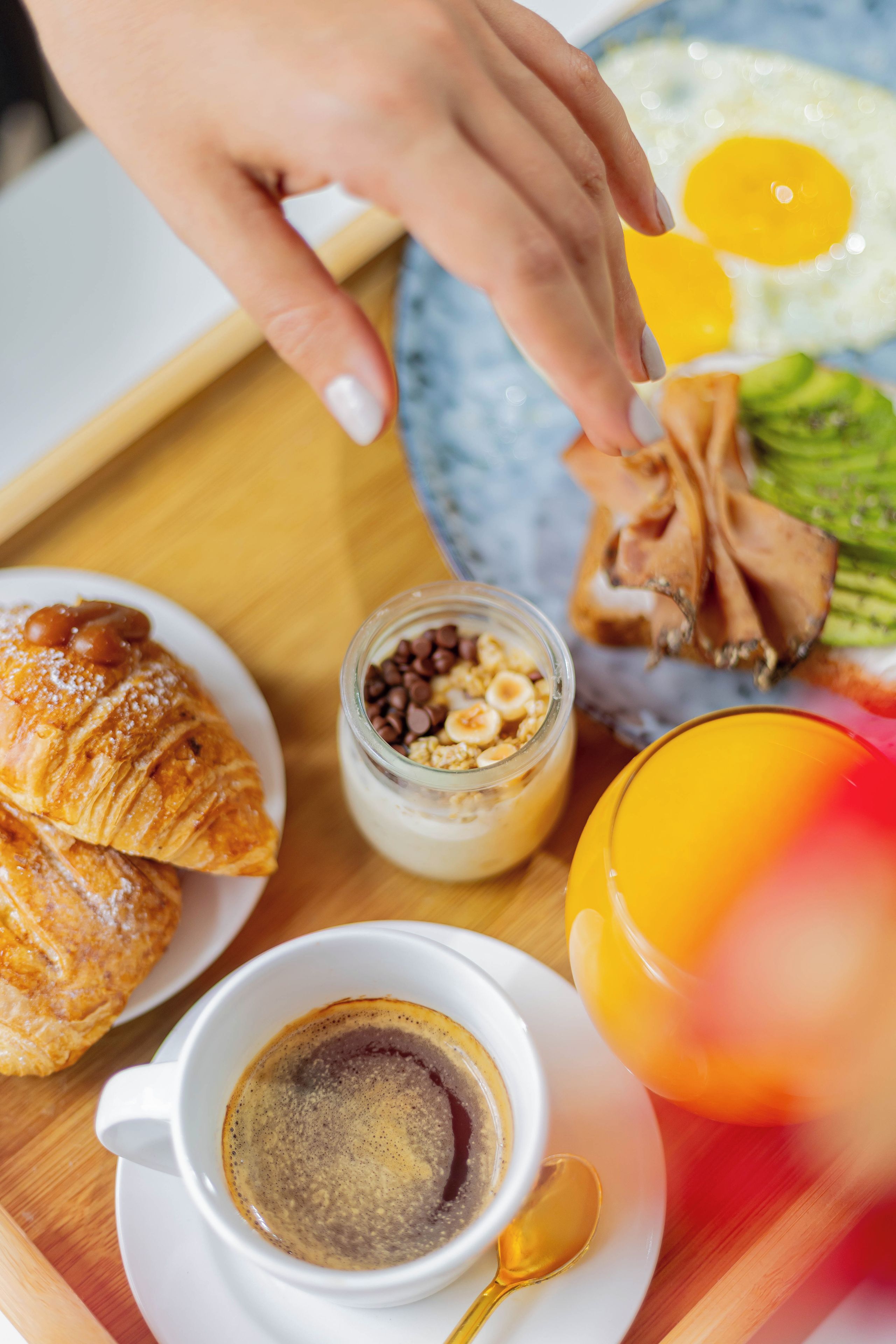 A breakfast setting with a croissant, a glass jar of granola, a plate with eggs, a cup of coffee, and a spoon.