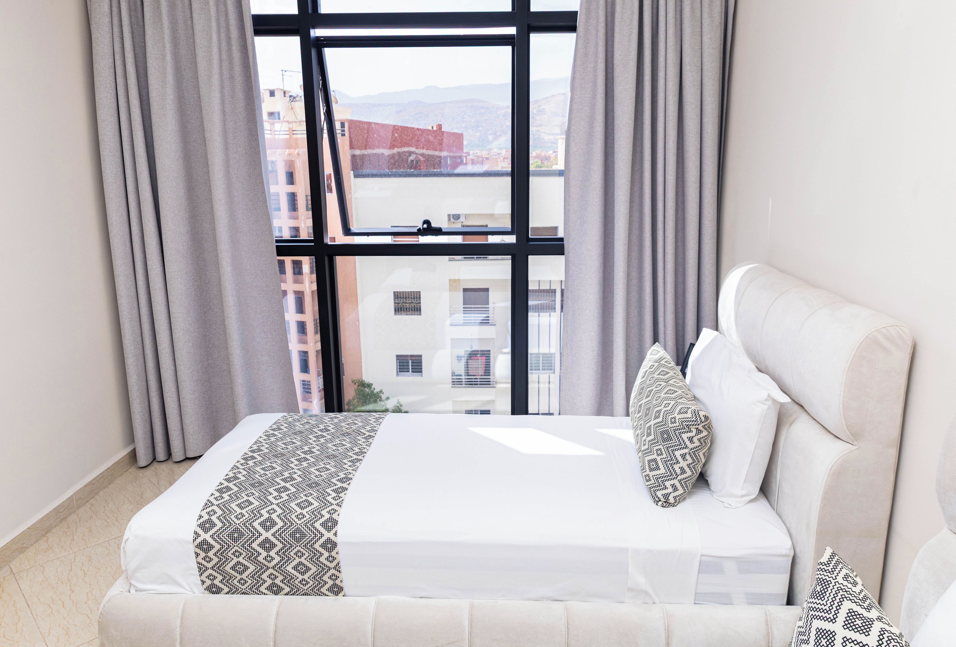 A hotel room with a large window, a bed with patterned pillows, and a view of buildings outside.