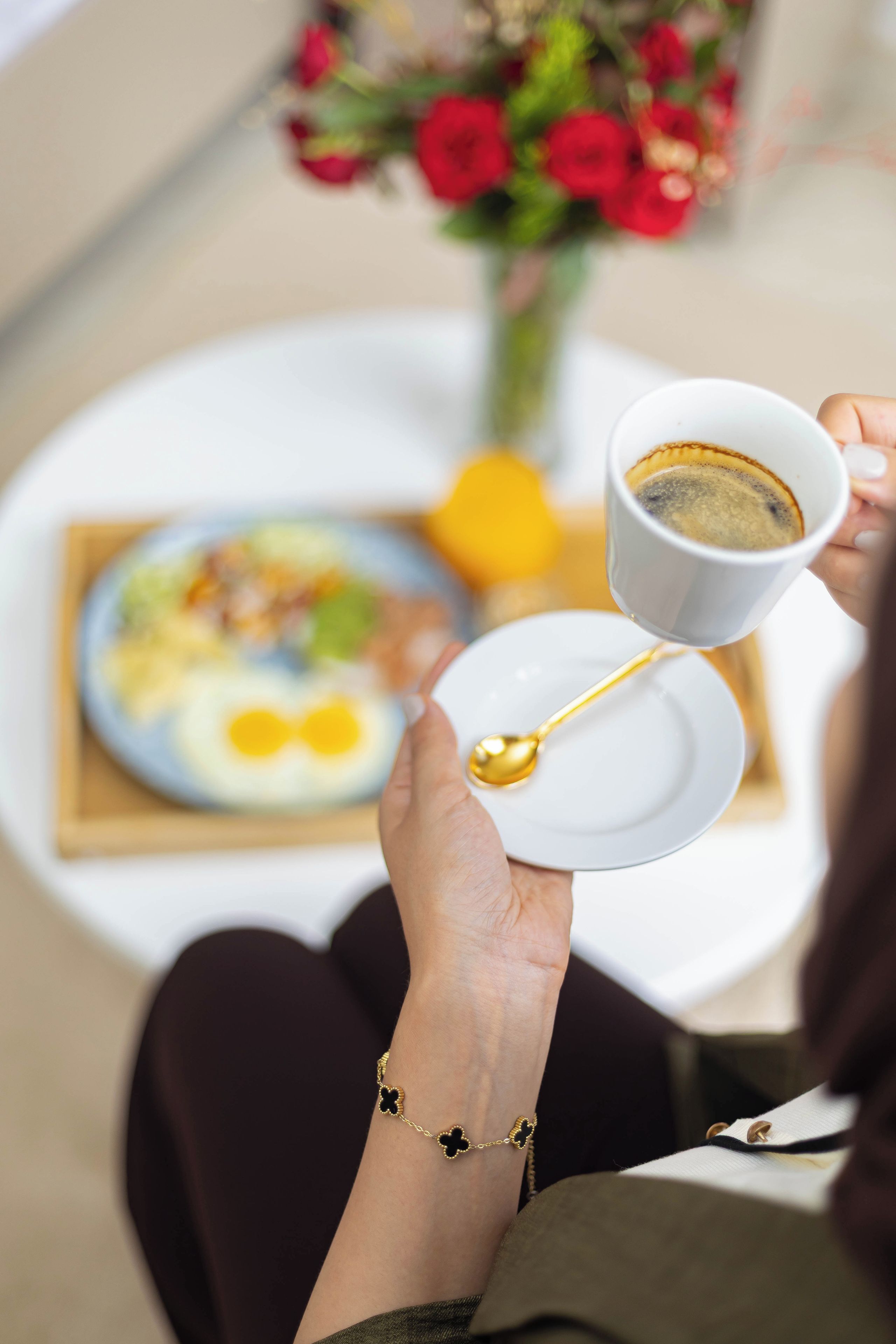 Une personne tenant une tasse de café avec une cuillère, assise à une table devant une assiette de nourriture, avec un vase de fleurs en arrière-plan.