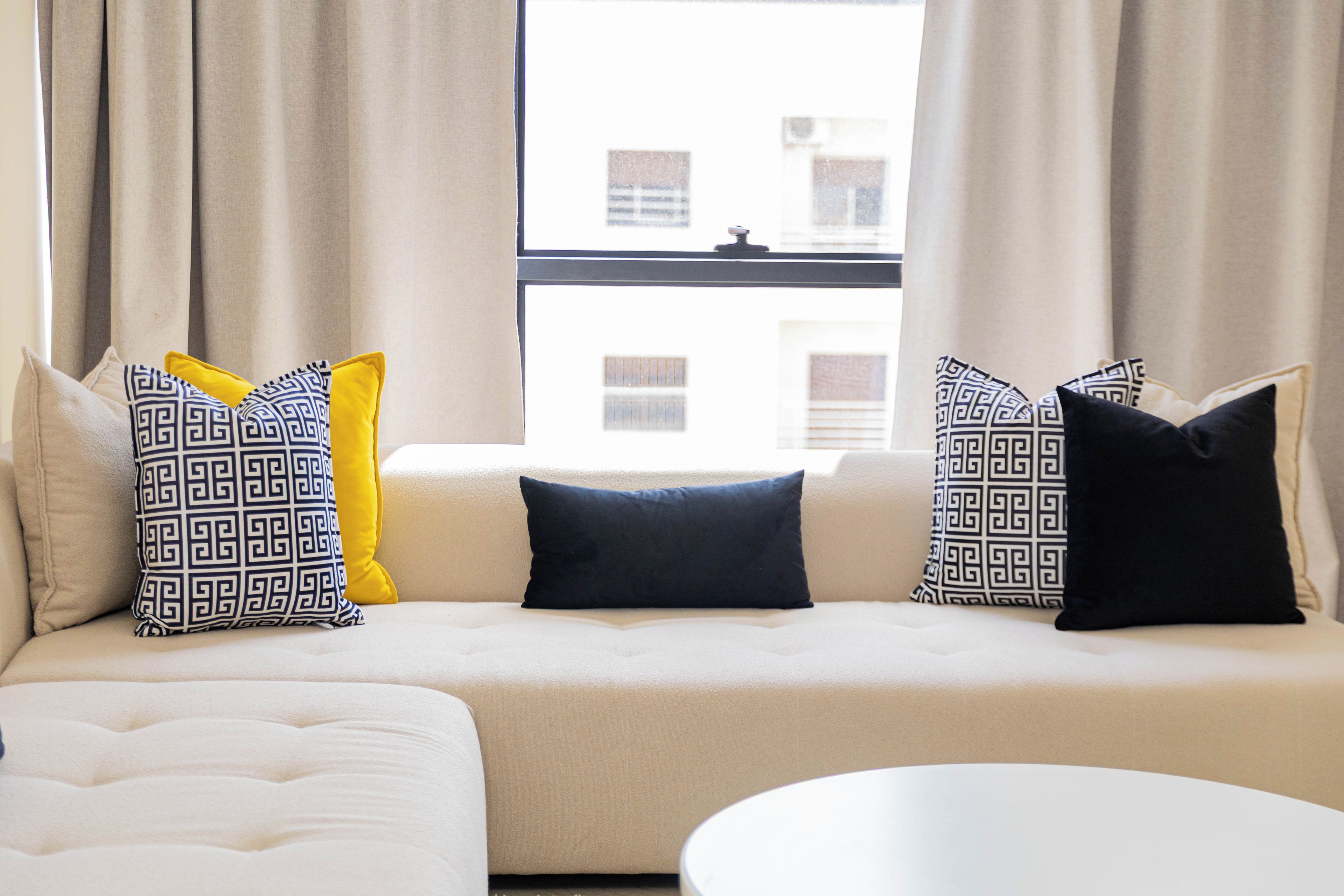 A modern living room with a beige sofa, patterned pillows, and a view of buildings through a window.