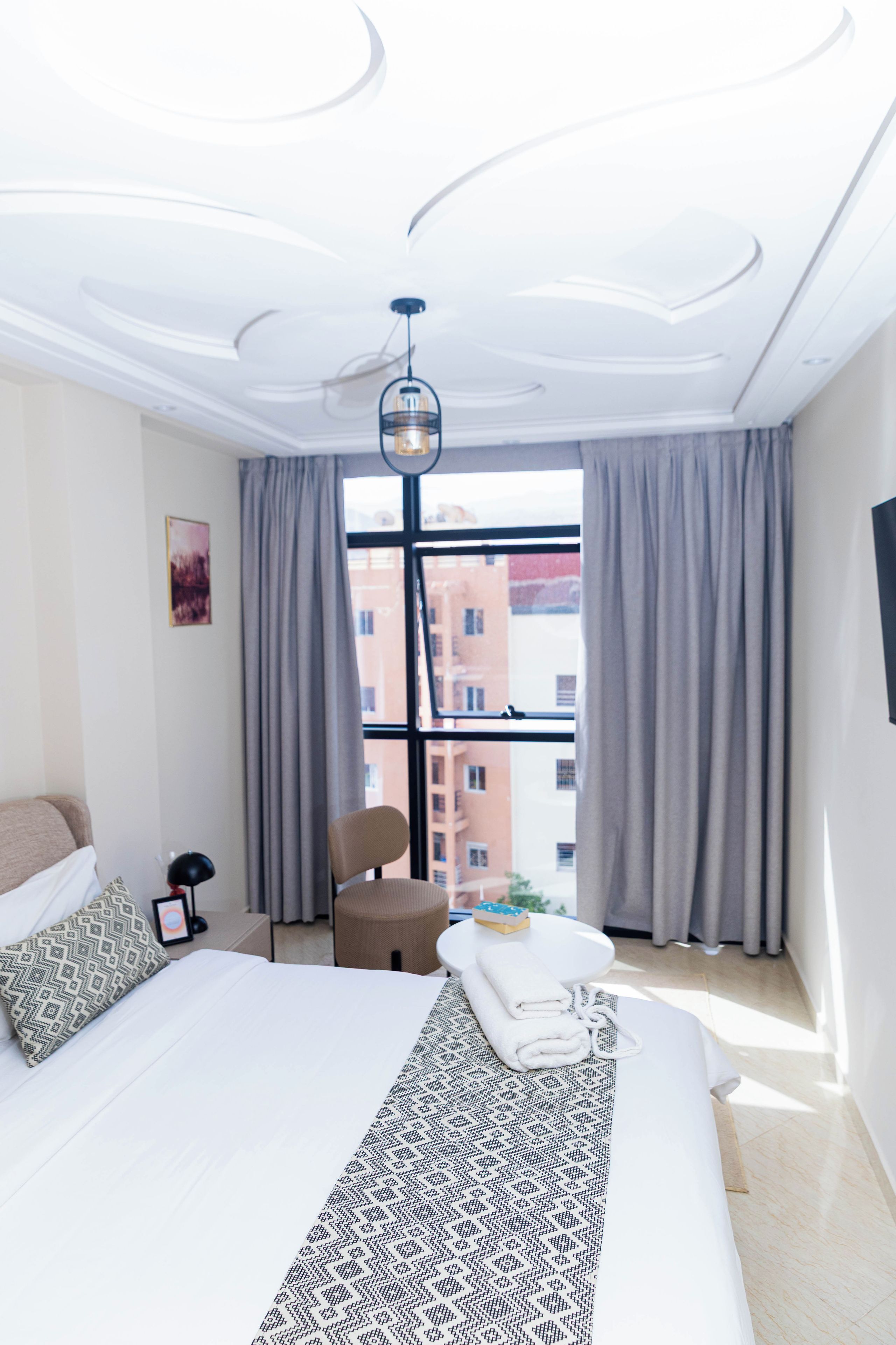 A modern hotel room with a large bed, a chair, a desk, and a view of buildings through large windows.
