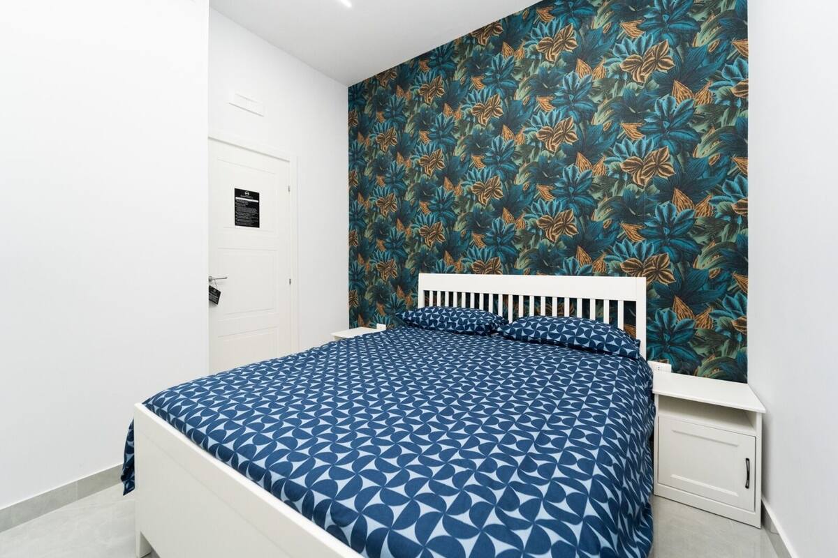 A bedroom with a bed, white headboard, and a wall with a floral pattern.
