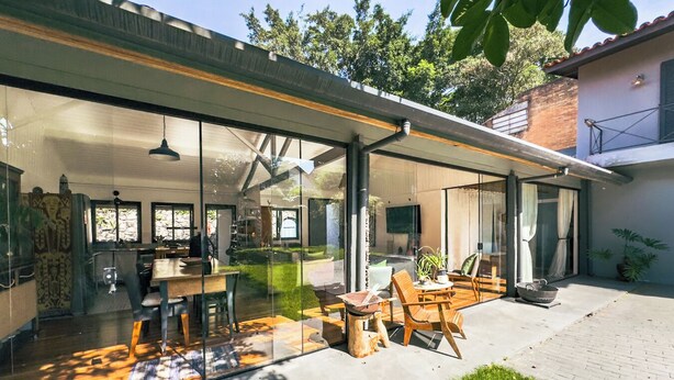 Ein modernes Haus mit einer gläsernen Veranda, Sitzgelegenheiten im Freien und einem sichtbaren Küchenbereich.