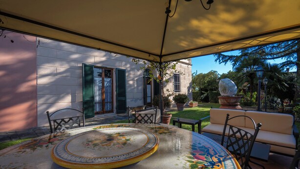 Una mesa redonda con mantel floral, un sofá beige y una planta en maceta en un espacio exterior cubierto, con un edificio al fondo.