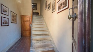 Un pasillo estrecho con una puerta de madera al final, una escalera con barandilla y varias imágenes enmarcadas en las paredes.