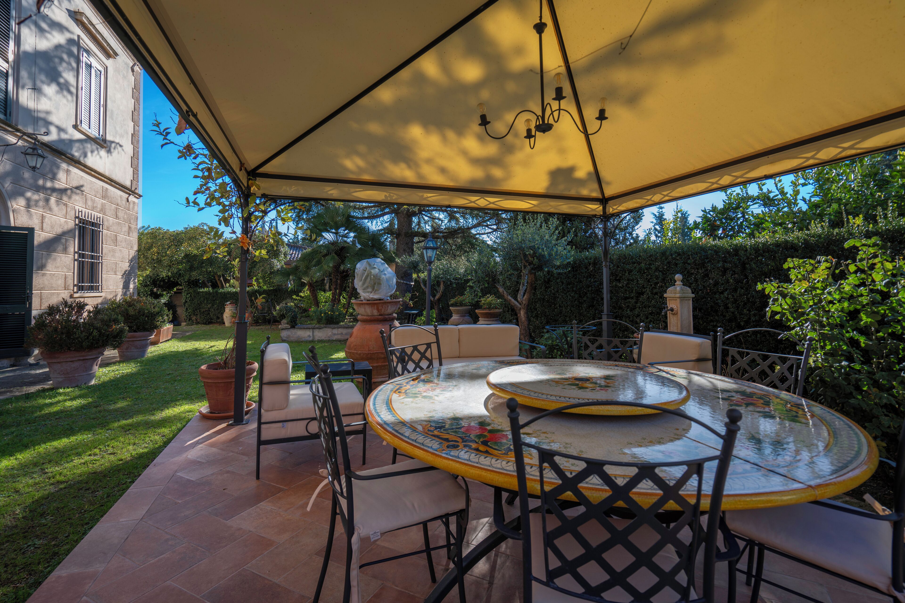 Une terrasse extérieure avec une table ronde et des chaises, entourée de plantes en pot et d’un bâtiment en arrière-plan.