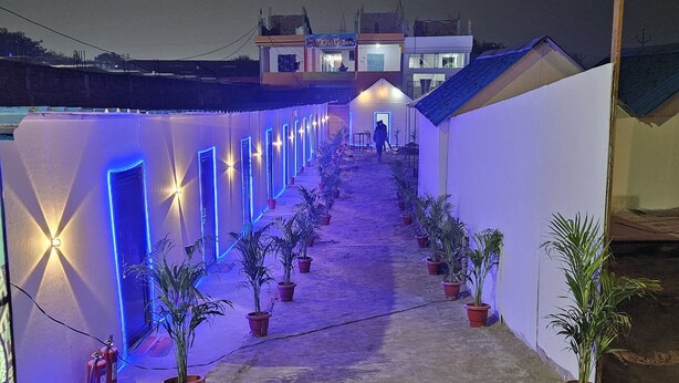 A corridor with potted plants and illuminated doors, leading to a building with a sign. - The Mahakumbh Vatika (Karchana)