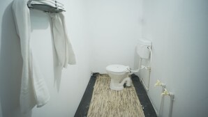 A small, white bathroom with a toilet, a towel rack, and a black and beige floor mat. - The Mahakumbh Vatika (Karchana)