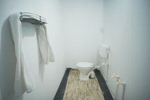 A small, white bathroom with a toilet, a towel rack, and a black and beige floor mat.