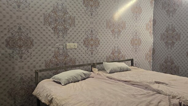 Een tweepersoonsbed met houten frame, twee kussens en een lichtgekleurd dekbed in een kamer met behang met patroon en een zichtbaar stopcontact aan de muur.