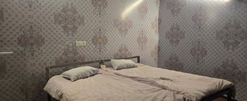 A double bed with a wooden frame, two pillows, and a light-colored bedspread in a room with patterned wallpaper and a visible electrical outlet on the wall.