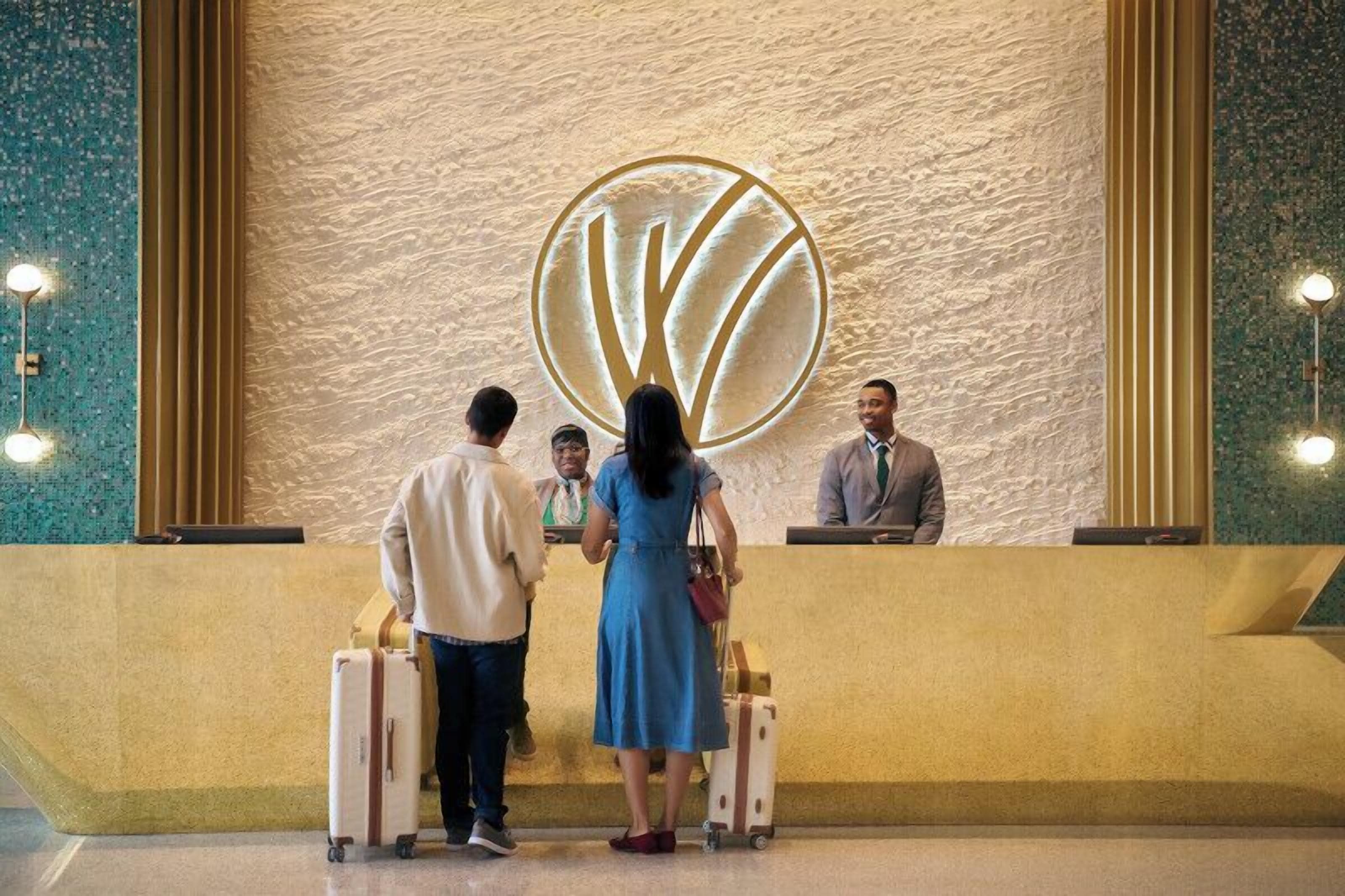 Una recepció amb un logotip gran, un home i una dona de peu, i una tercera persona asseguda darrere del taulell.